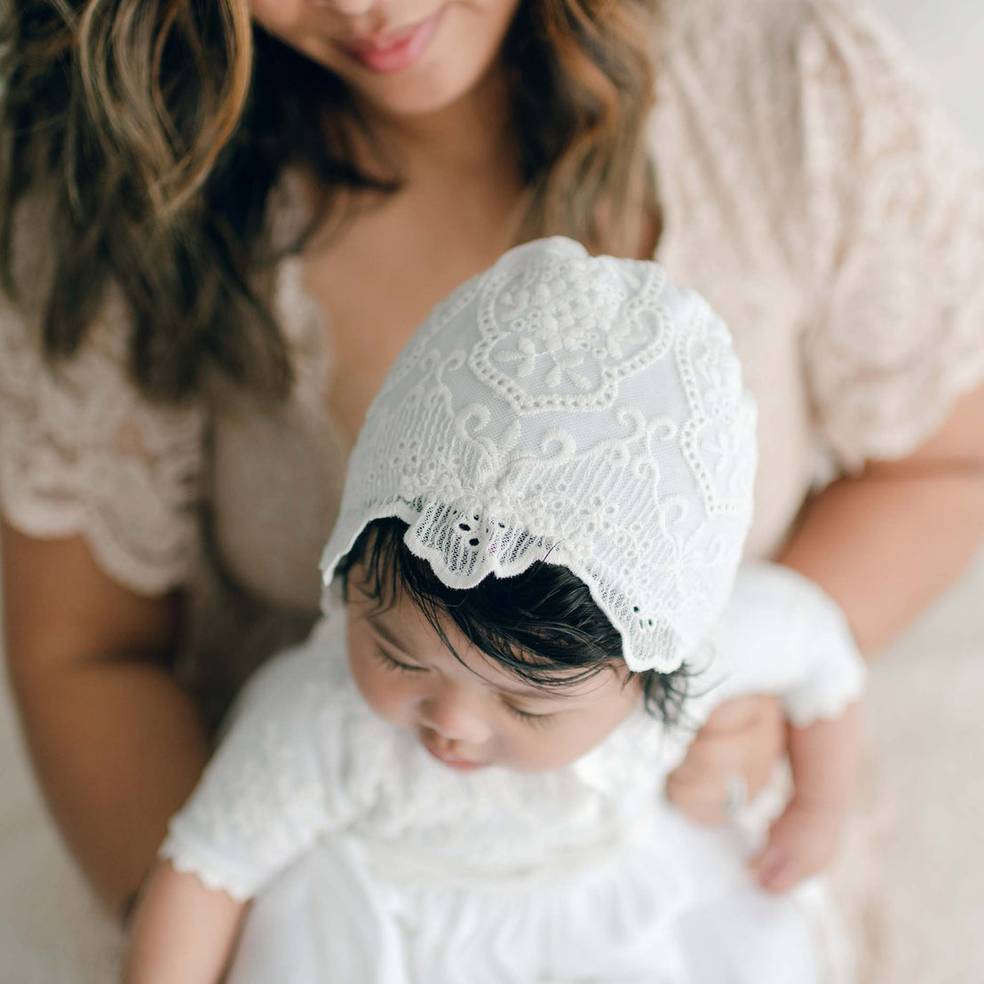 A woman with wavy hair, partially visible, holds a baby dressed in white. The baby, adorned in a delicate handmade Eliza Bonnet, looks downward. The background is softly blurred.