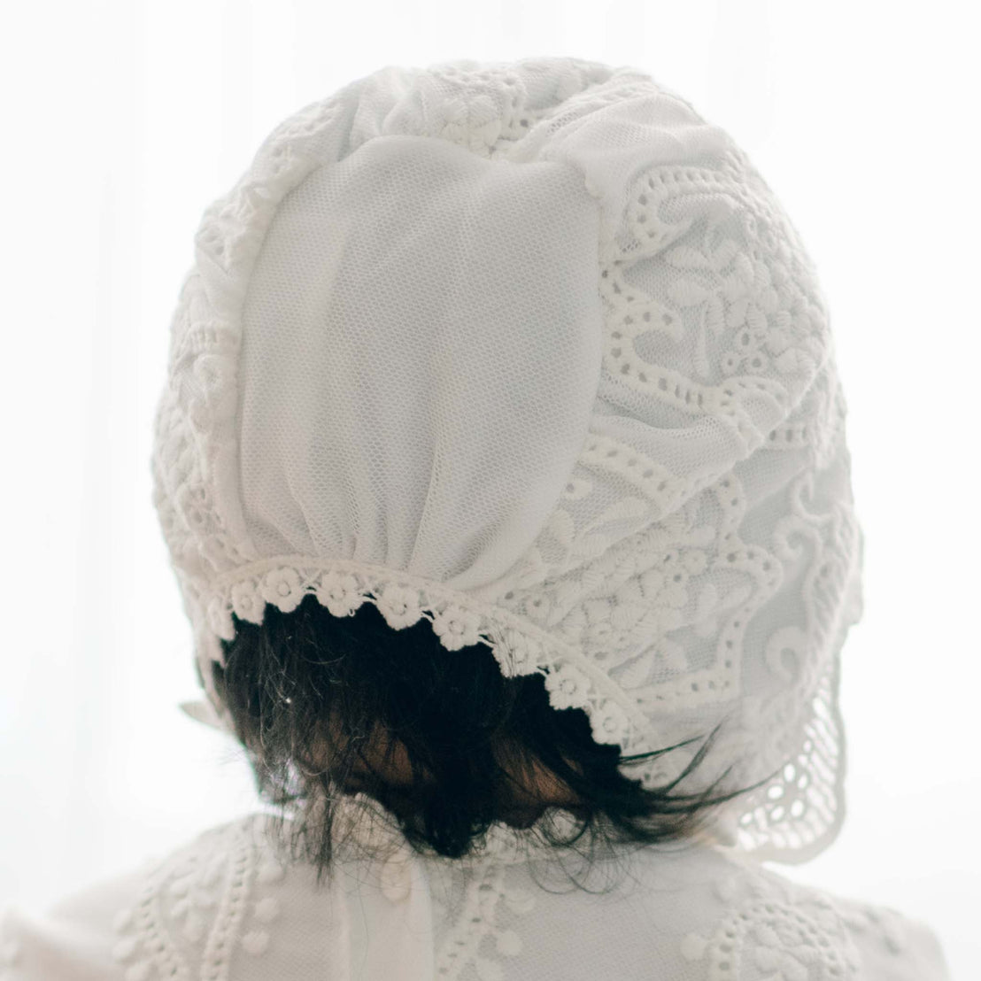 A close-up of the back of a baby's head, showing intricate lacework on a handmade white Eliza Bonnet. The bonnet, made from soft pima cotton with detailed embroidery, covers most of the head, with some dark hair visible at the neck. The baby is wearing a matching white garment. The background is softly lit.