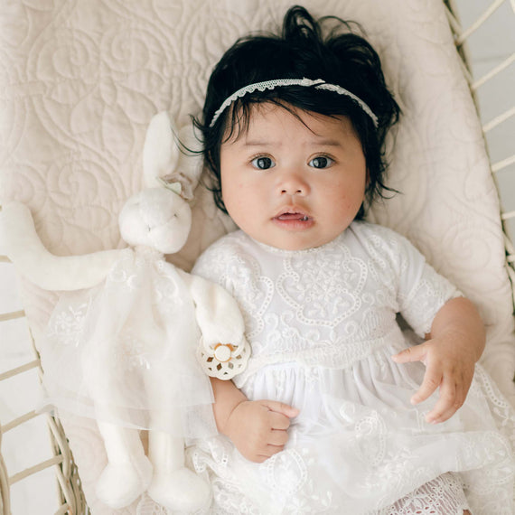 An infant with dark hair lies on a quilted, off-white surface, wearing a delicate white lace dress and a lace headband. The baby holds an Eliza Silly Bunny Buddy | Pacifier Holder dressed in matching lace and has a pacifier clip attached to its garment. The baby's wide eyes look directly at the camera.