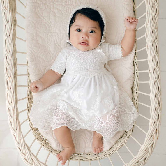 A baby wearing an Eliza Lace Dress & Headband is lying in a woven bassinet. The baby has dark hair and wide eyes, looking up with one arm raised. The bed has a quilted white mattress, and the background is soft and neutral in color.