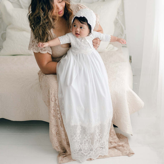 A woman in a lace dress sits on a bed holding a baby dressed in an Eliza Blessing Gown & Bonnet. They both have dark curly hair. The room has a light and airy feel with white decor and a white background, enhancing the elegance of their christening accessories, all handmade in the USA.