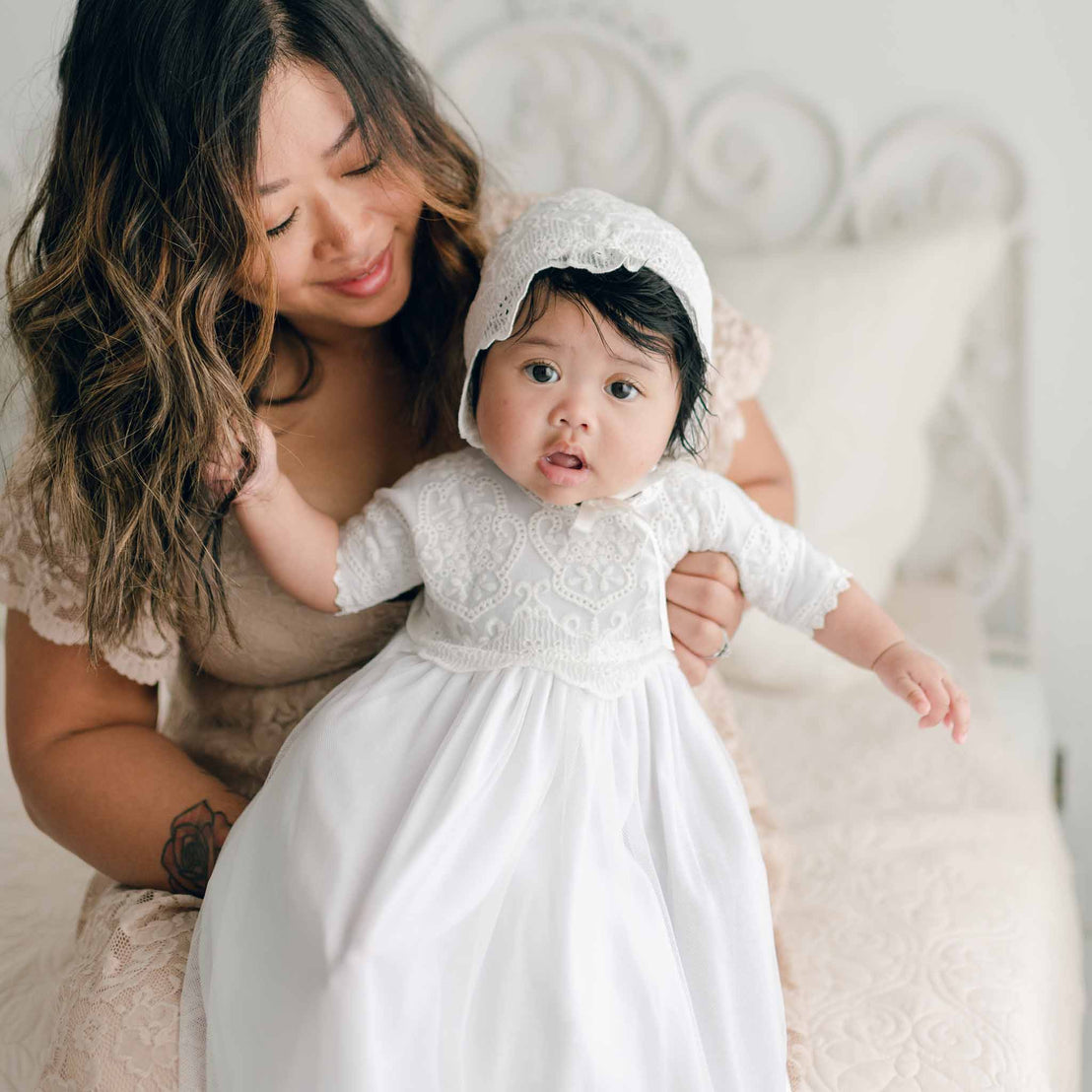 A woman with long, wavy hair smiles while holding a baby dressed in an Eliza Blessing Gown & Bonnet. They are sitting on a white bed in a brightly lit room. The baby, wearing these exquisite Christening accessories handmade in the USA, has a curious expression and reaches out with one hand.