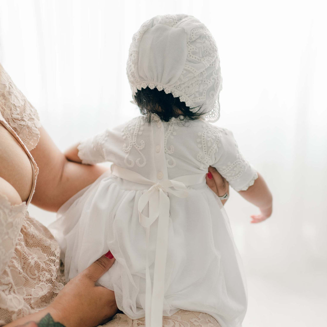 A baby in a white Eliza Blessing Gown & Bonnet is being held from behind by an adult dressed in lace. The photo is softly lit, creating a serene and gentle atmosphere. The baby, adorned with Christening accessories, is looking away from the camera.