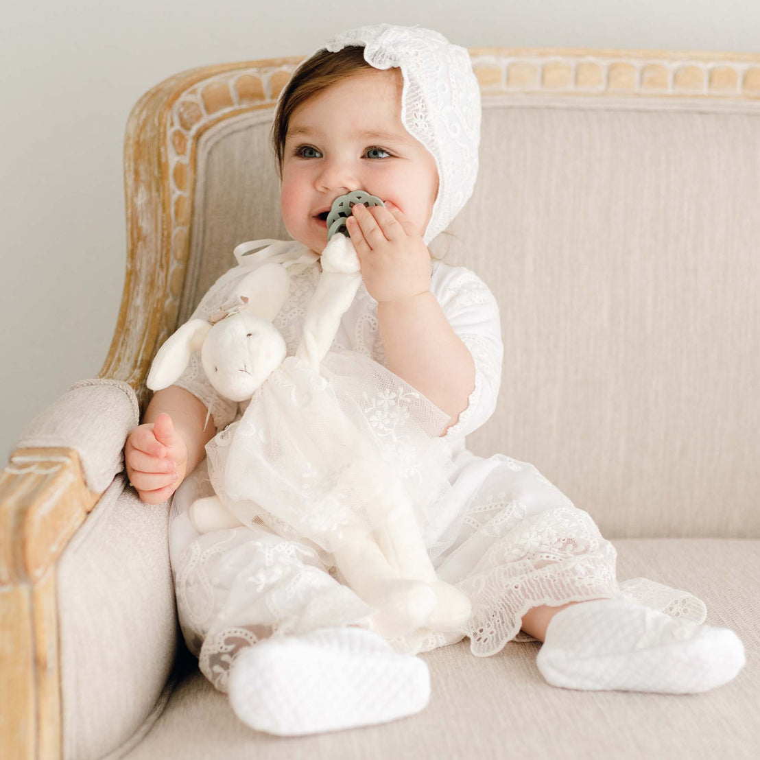 A joyful baby in a white lace heirloom outfit sitting on a beige upscale sofa, playfully biting an Eliza Silly Bunny Buddy | Pacifier Holder.