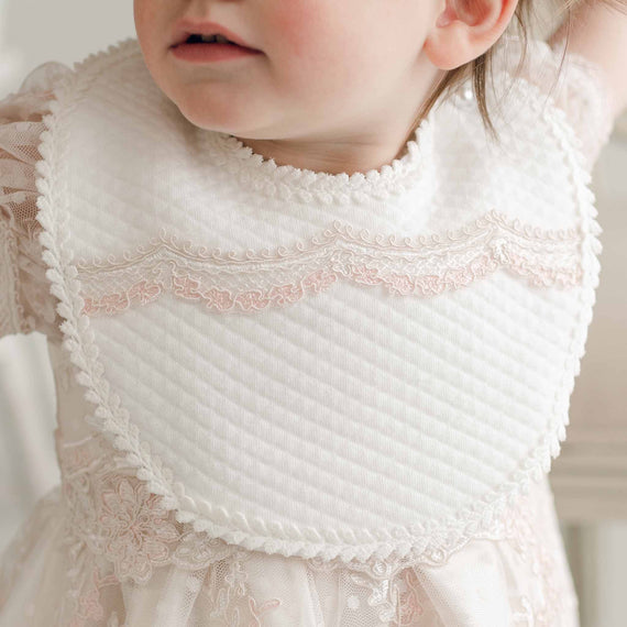 Close-up of a young child wearing an Elizabeth Bib, a white, textured baby bib with delicate pink lace trim. The child is dressed in a white baptism outfit adorned with matching pink lace patterns. Only the lower half of the child's face and upper torso are visible, capturing the elegance of the moment.