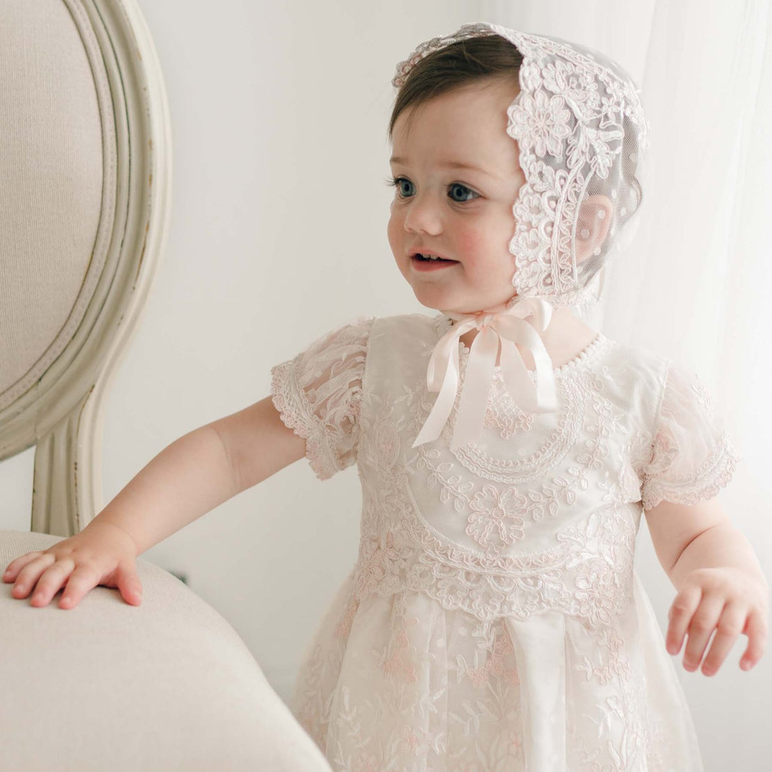 A toddler wearing the delicate Elizabeth Christening Dress in light pink, featuring intricate embroidered lace patterns and a matching bonnet, stands beside a cream-colored upholstered chair, gazing off to the side, smiling. The room has a soft, neutral decor with light streaming through a window.