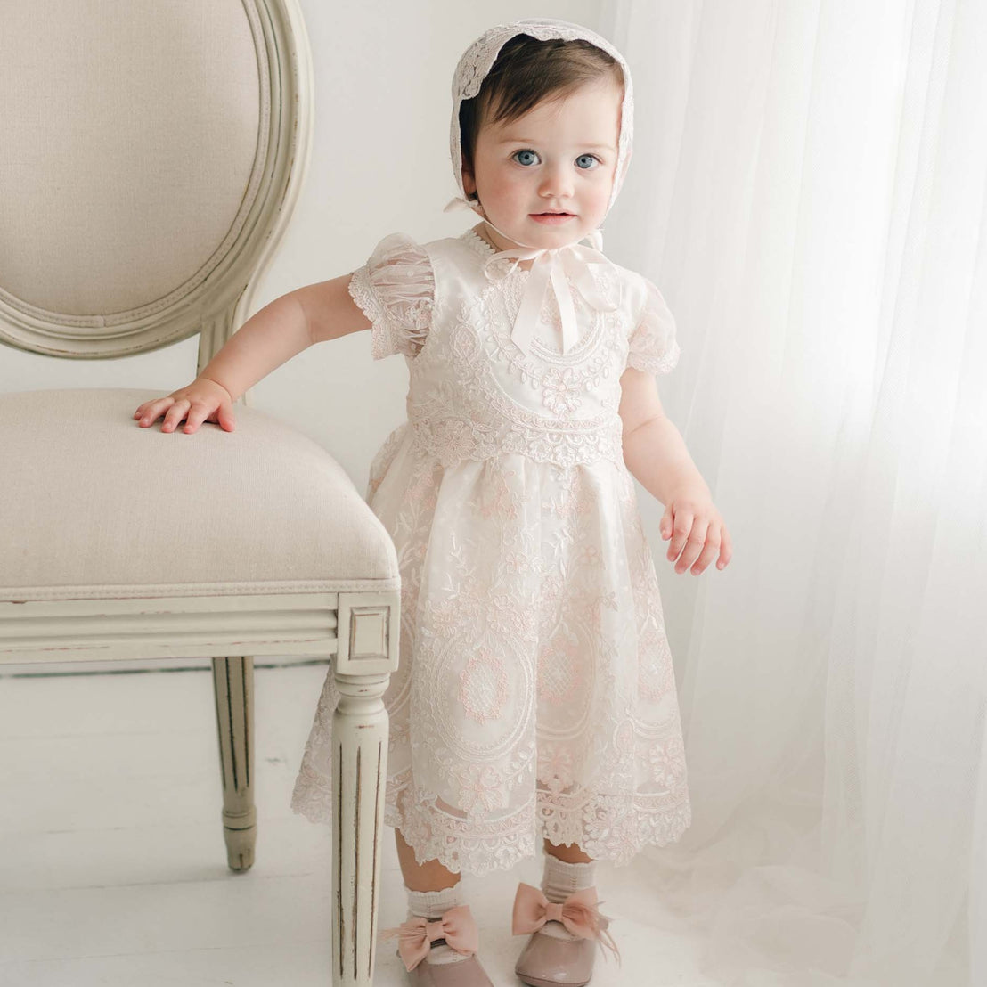 A toddler wearing the delicate, handmade Elizabeth Christening Dress with embroidered lace and a matching bonnet stands beside a vintage-style upholstered chair. The child has one hand resting on the chair and is looking slightly to the side, with soft natural light filtering through a sheer curtain in the background.