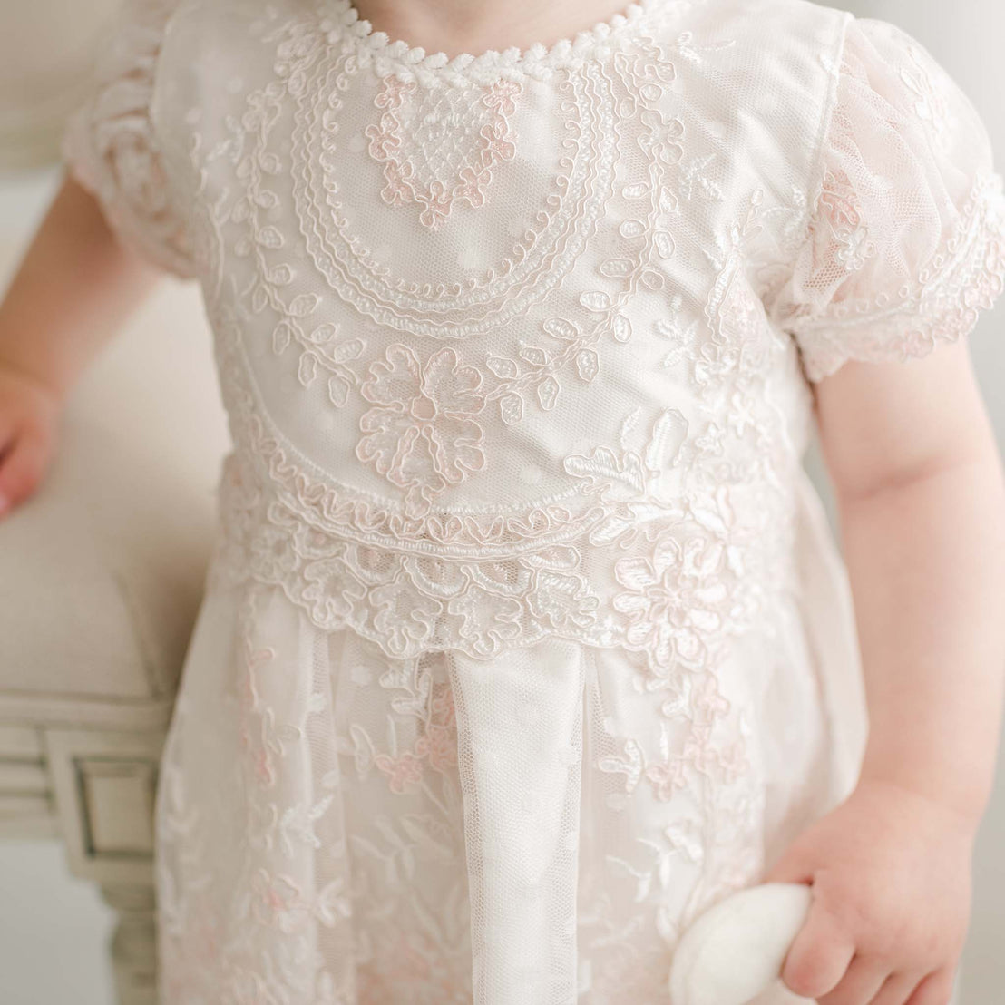 A close-up of a young child wearing the Elizabeth Christening Dress, adorned with delicate floral embroidery and intricate lace. The child is holding a small white object in one hand while standing next to a cushioned piece of furniture. The image beautifully captures the handcrafted details of the dress.