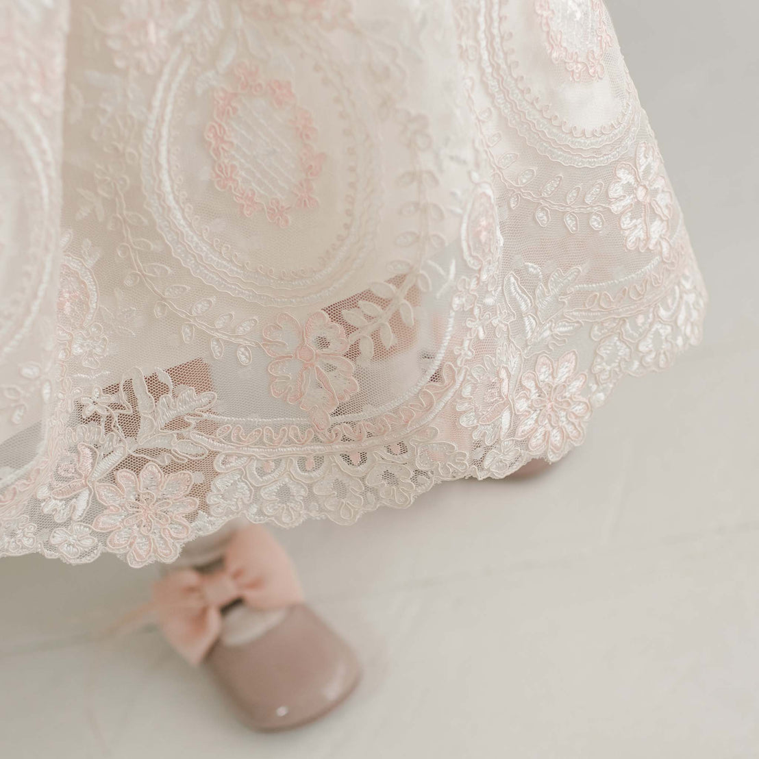 Close-up of the hem of the delicate Elizabeth Christening Dress in white and pink, featuring intricate floral patterns in embroidered lace. The dress is paired with shiny, blush-colored shoes adorned with pink bows. The background is a light, neutral color.