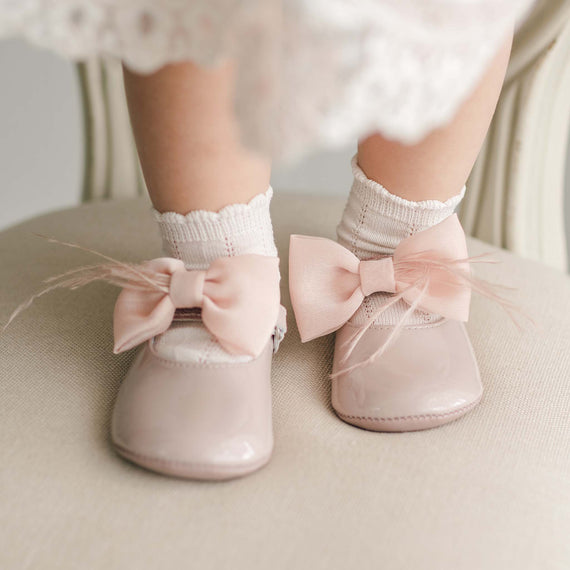 Close-up a baby's feet wearing the Rose Patent Leather Mary Janes. They are made with pink patent leather with a soft velcro strap that features a rose pink chiffon bow and feather. The child is also wearing white lace-trimmed socks and the background shows a blurred portion of a beige chair's upholstery.