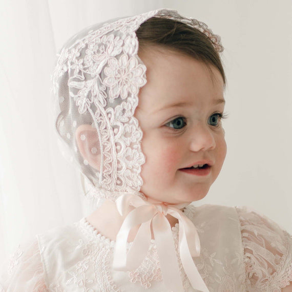 A young child with fair skin and dark hair adorns the Elizabeth Lace Bonnet, a delicate light pink lace piece tied with a silk ribbon under the chin. The handmade bonnet features intricate embroidery, perfectly complementing the matching lace garment. Against a soft white background, the child looks off to the side with a slight smile.