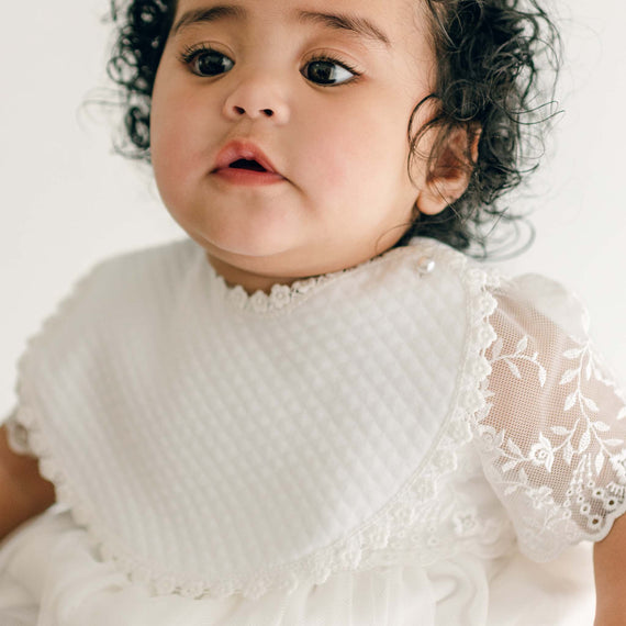 A baby girl with curly hair and expressive eyes wears the Ella Bib and Romper Dress, looking slightly off-camera.