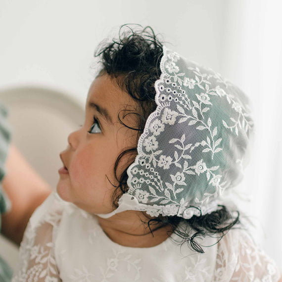 A baby with dark curly hair wearing the Ella Lace Bonnet and matching Ella baptism gown glances sideways.