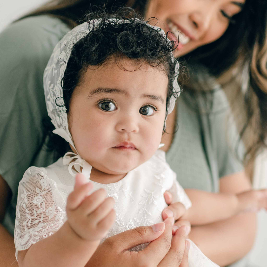 A baby with curly hair and large eyes, wearing the Ella baptism bonnet, and held by her mother.