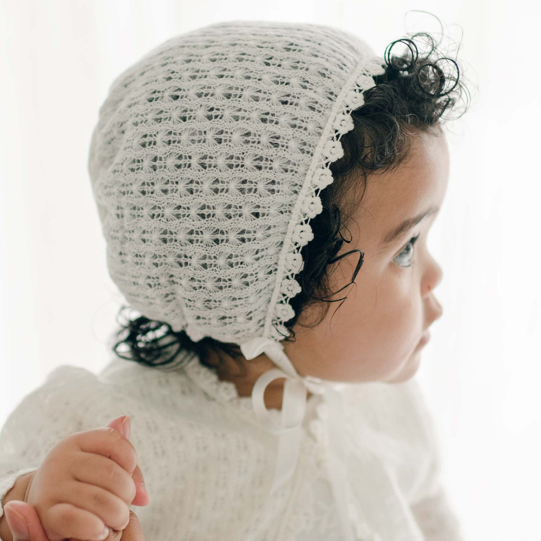 A young child with curly hair is wearing a white, lacy bonnet and matching Eliza Knit Sweater made of soft cotton lace. The child is facing to the side and holding an adult's hand, gazing intently at something off-camera. The background is bright and softly lit.