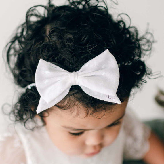 A close-up photo of a toddler with dark curly hair adorned with a white Ella Bow Headband, looking down thoughtfully.