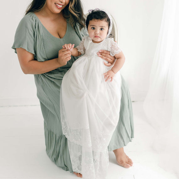 A baby girl in the Ella Christening Gown and Ella Lace Bonnet, held by her mother.