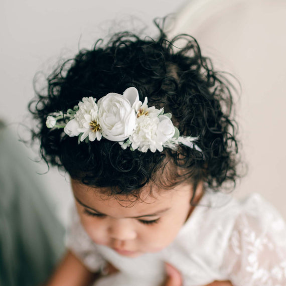 A baby with curly dark hair gazes downward while wearing the boutique Ella Flower Headband, adorned with delicate white flowers.