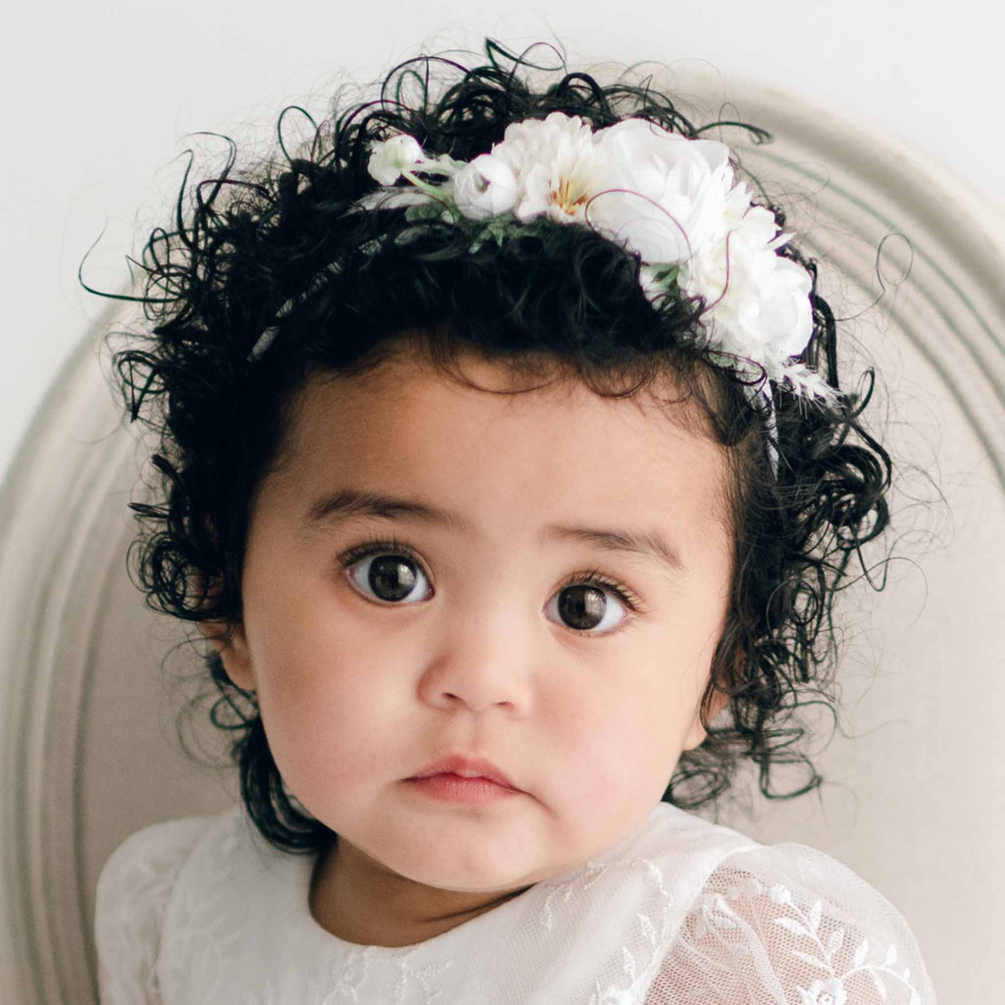 A baby with curly dark hair adorned with the Ella Flower Headband, featuring white flowers, looks directly at the camera.