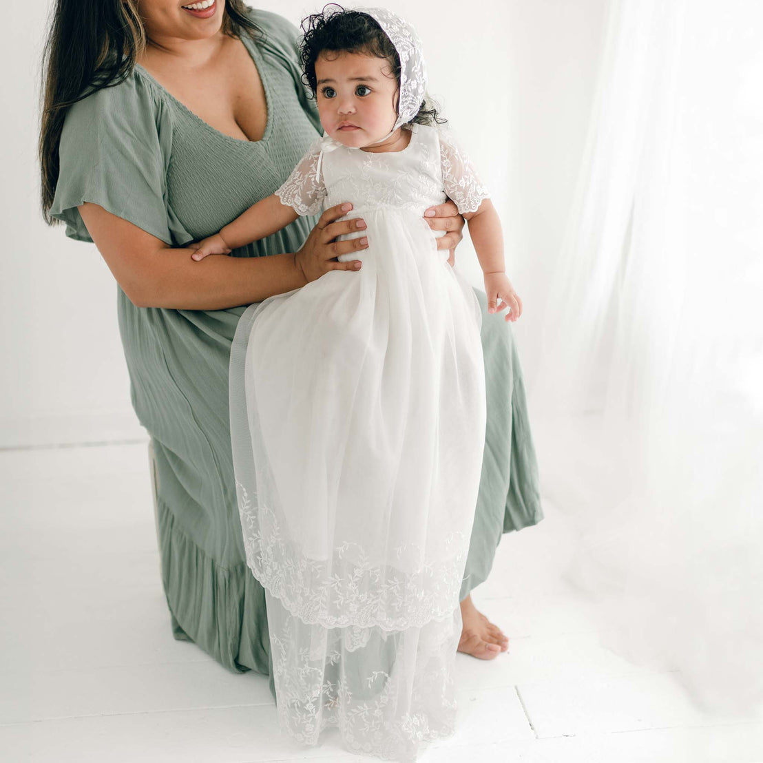 A woman in a green dress holds her baby daughter dressed in the Ella Christening Gown.