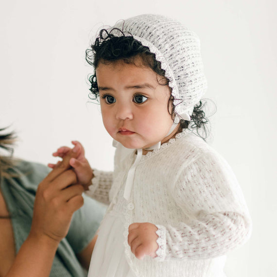 A baby girl with dark curly hair wears an Ella Knit Bonnet and matching Ella Knit Sweater, looking thoughtfully off to the side.