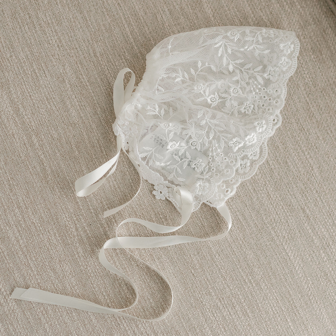 A flat lay photo of the Ella lace christening bonnet on a beige fabric chair.