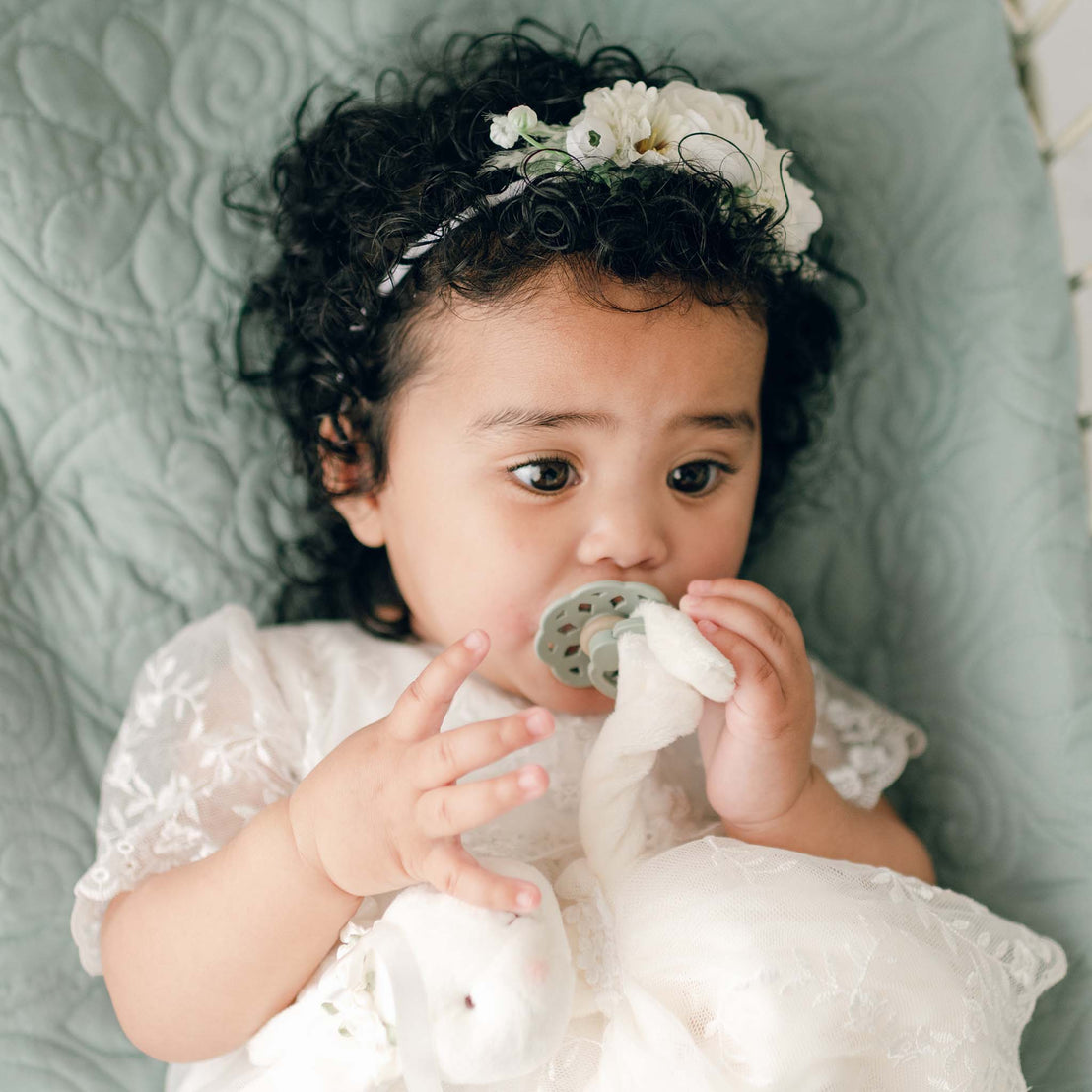 A baby with curly hair, wearing the Ella Flower Headband, lies on a quilted surface and holds an Ella Pacifier in sage and an Ella Silly Bunny Buddy.