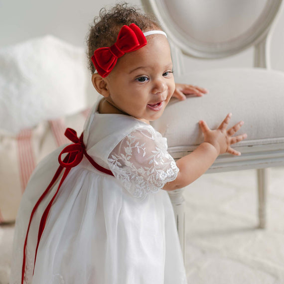 A baby wearing an Ella Holiday Romper Dress, characterized by its vintage-style design featuring embroidered netting lace and lace sleeves, stands holding a white chair. The baby completes the charming look with a red bow headband and smiles against a light-colored background.