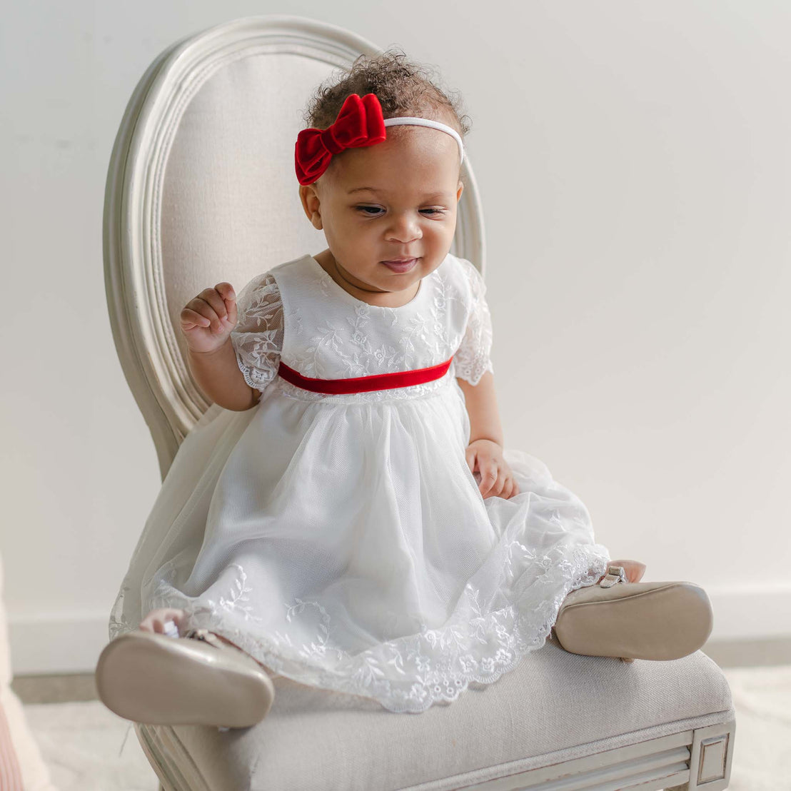 A baby in the Ella Holiday Romper Dress, featuring delicate floral embroidered lace and a red velvet ribbon sash, sits on an elegant chair. The baby wears a headband adorned with a red bow and beige shoes, delighting with a playful expression as one eye is slightly squinted.