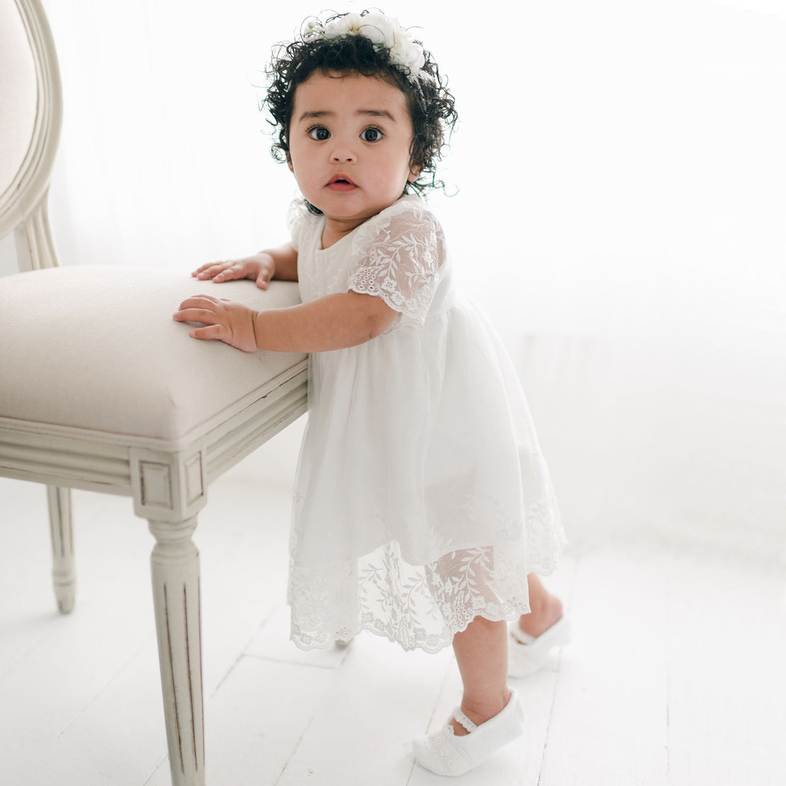 A toddler with curly hair, wearing a boutique Ella baptism dress and matching booties, stands and holds onto a cream-colored upholstered chair in a brightly lit room.