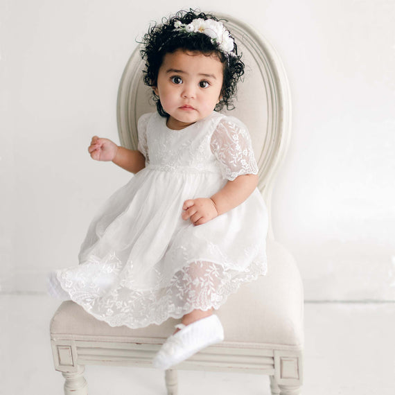 A baby girl with curly hair, wearing the Ella baptism dress flower headband, sits on an light beige chair and looks forward with a curious expression.