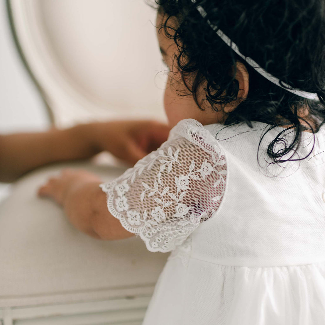 Viewed from behind, a toddler with dark curly hair wears an vintage style Ella romper dress and holds onto a upholstered chair.