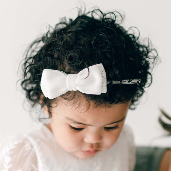 A baby girl with dark curly hair wears the Ella Velvet Bow Headband, looking down away from the camera.