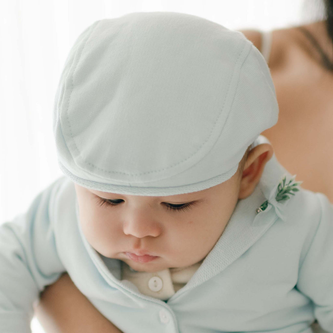 A close-up of a baby wearing an Ezra Powder Blue Newsboy Cap and matching outfit. The baby is looking down with a calm expression, embodying a classic Christening look. The background is softly lit, and a woman's shoulder is partially visible on the right side of the image.