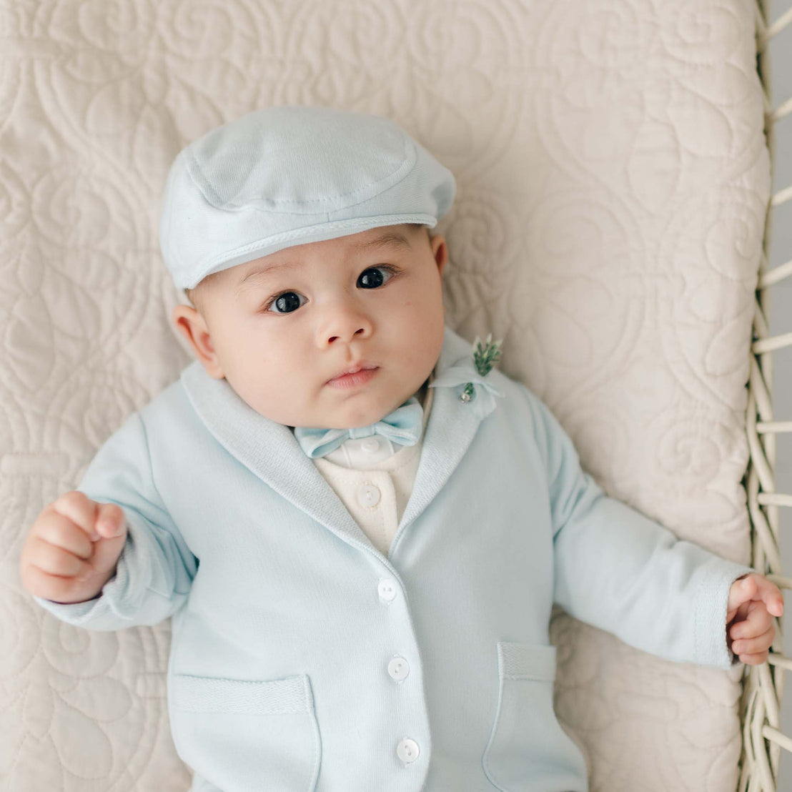 A baby, dressed in an Ezra Powder Blue 4-Piece Suit made of soft French Terry cotton, is lying down on a quilted surface. The outfit, handmade in the USA, includes a matching light blue flat cap, bow tie, and cardigan with buttons. The baby looks at the camera with a calm expression, hands slightly raised.