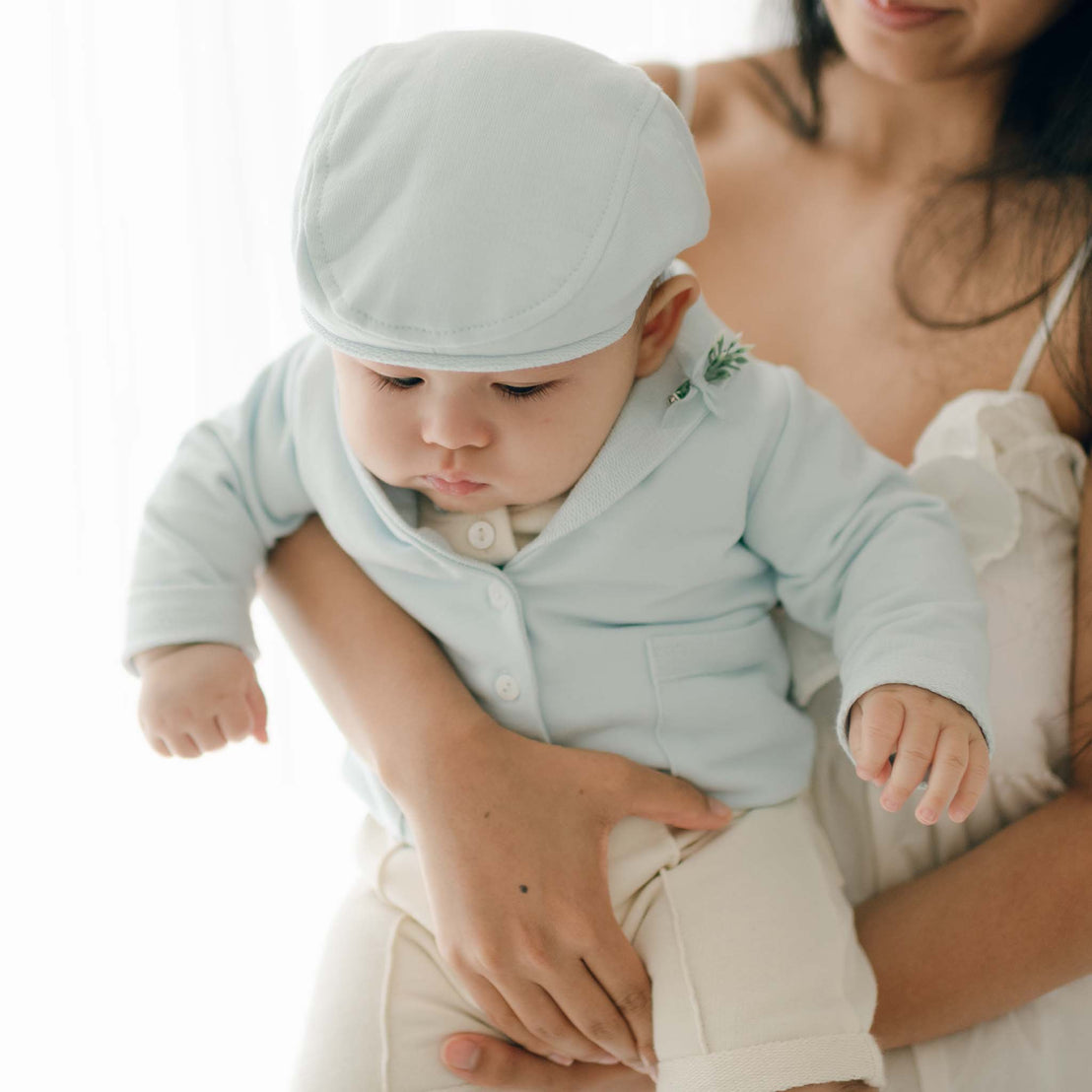 A baby dressed in an Ezra Powder Blue 4-Piece Suit made of light blue French Terry cotton is being held by a woman wearing a white, sleeveless dress. The baby looks downward, and the woman's face is mostly out of the frame. The background is bright and softly lit.