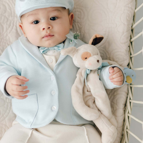 A baby dressed in a pale blue jacket, matching hat, and bow tie is lying on a quilted blanket. The baby is holding an Ezra Powder Blue Silly Puppy Buddy | Pacifier Holder. A pacifier clip dangles nearby, completing the calm and cozy scene.