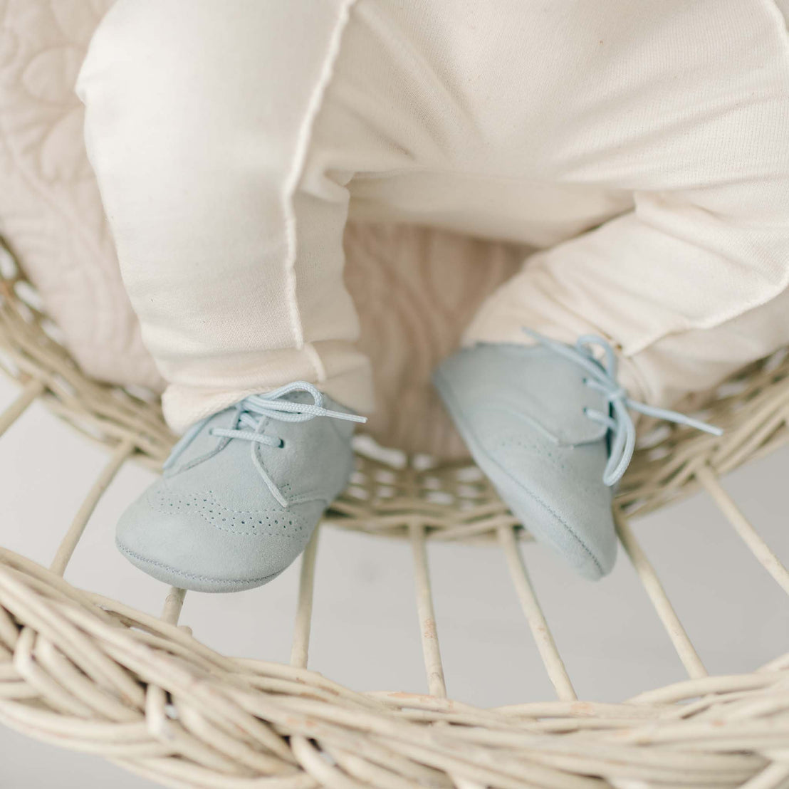 A baby wearing Ezra Suede Shoes and cream-colored pants is seated on a white, woven wicker chair. The focus is on the baby's feet and shoes, with soft, neutral tones dominating the image.