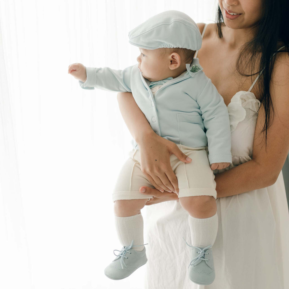 A woman holding a baby dressed in an Ezra Powder Blue 4-Piece Suit, featuring a light blue cap, jacket, white shorts, and knee-high socks. The woman is wearing a white dress. They are standing in front of a sheer white curtain. The baby is reaching out with one hand.