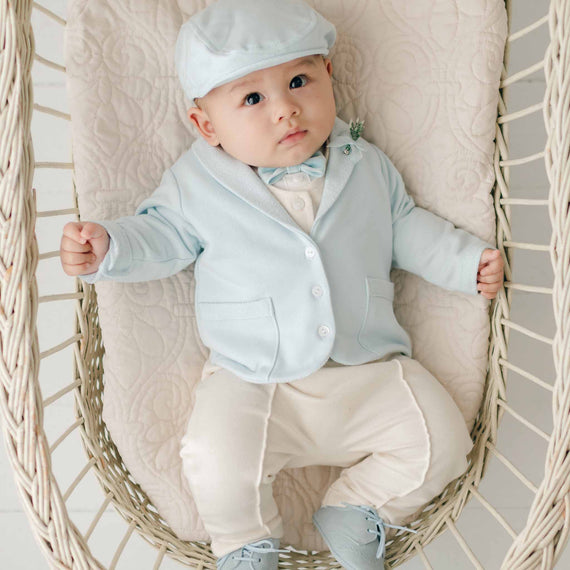 A baby dressed in an Ezra Powder Blue 4-Piece Suit with a light blue cap, matching jacket made from French Terry cotton, and beige pants sits in a wicker chair. The baby has a curious expression, with hands holding onto the chair's sides and feet in light blue shoes, resting on a beige cushioned seat.