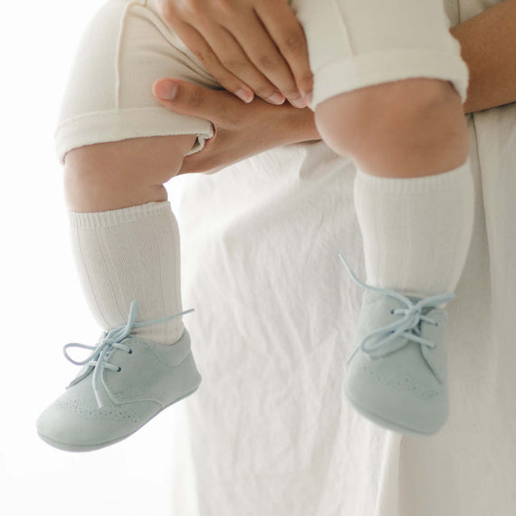 A person is holding a baby dressed in white pants and Cream Ribbed Socks. The baby is wearing light blue lace-up shoes. The baby is photographed from the waist down, with their legs and feet prominently displayed.