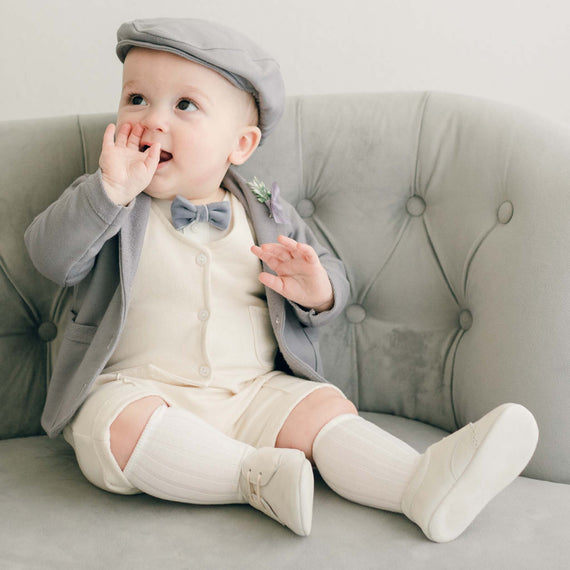 Baby boy sitting on a couch wearing the Ezra 4-Piece Suit with matching Newsboy Cap, heather velvet bow tie and boutonniere