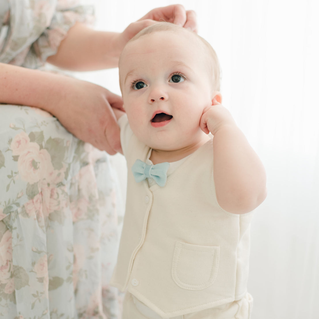 Baby boy wearing a tan vest with matching tan shorts. He is also wearing a powder blue velvet bow tie