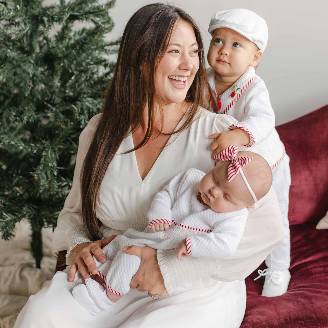 A woman smiles while sitting next to a Christmas tree, holding a sleeping baby dressed in the Nicholas 3-Piece Suit, featuring quilted cotton with red accents. A toddler in a matching outfit sits on her lap, wearing a white hat and looking up. They're all seated on a red couch.