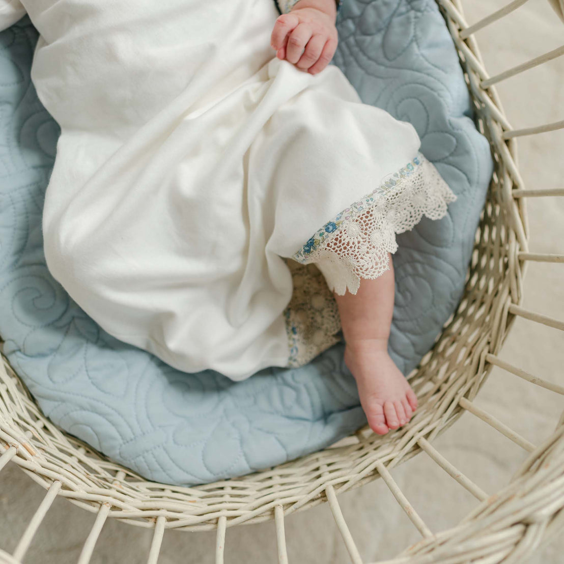 Close-up of a baby's leg resting in a basket, showing the scalloped lace trim of the Petite Fleur Layette gown.