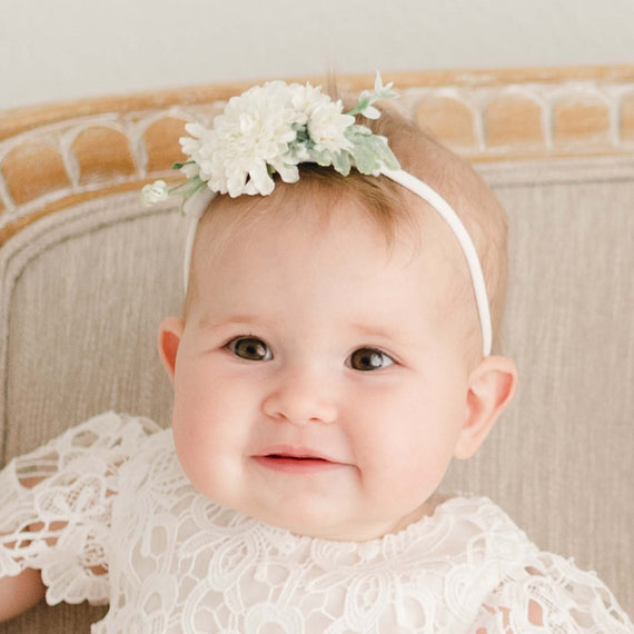 Baby girl wearing the Lola Flower Headband, featuring realistic cream flowers and soft green leaves, paired with a lace christening outfit.