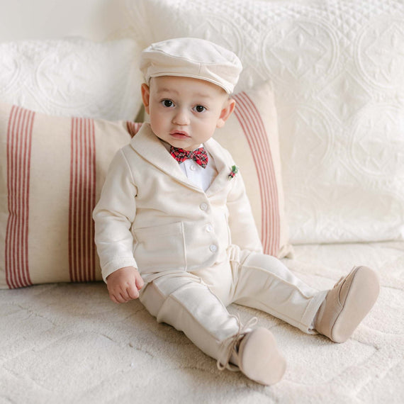 A baby wearing the Gabriel Suit & Bow Tie Set sits on a bed with striped pillows. The cream-colored, handmade suit comes with a matching cap and red bow tie, crafted from soft French Terry cotton. It's an ideal outfit for any holiday party as the baby looks curiously at the camera against off-white, textured bedding.