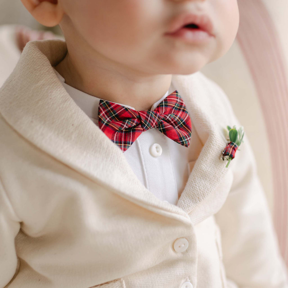 A close-up of a young child in a Gabriel 3-Piece Suit, featuring a cream-colored buttoned jacket, white shirt, and red plaid bow tie. A small boutonniere is placed on the jacket, with the child's face partially visible.