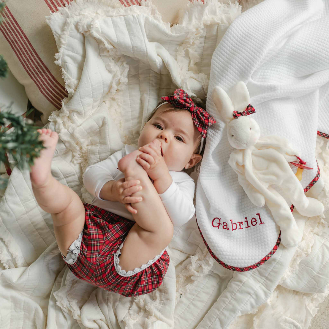 A baby in a white top and plaid shorts lies on a soft, textured blanket. The baby is playfully holding a foot with their hands. Beside them is the Gabriel Personalized Blanket and a plush bunny toy.