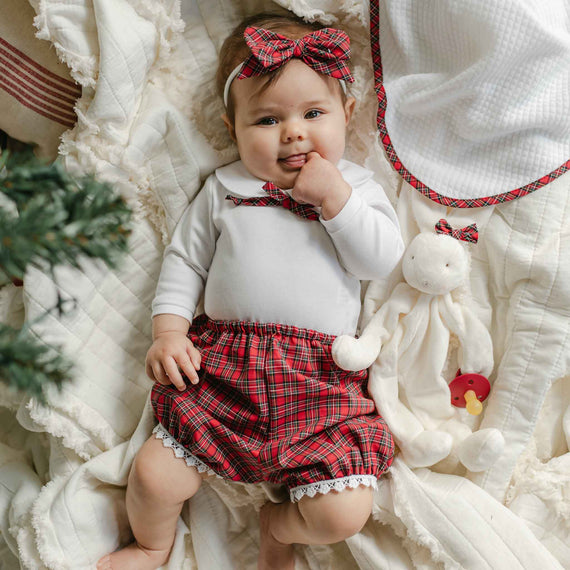 A baby in the Gabriella Bloomer Set, featuring a white cotton onesie and red plaid skirt with a coordinated headband, lies on a soft blanket. Accompanied by a plush toy and cloth, the baby has a hand near their mouth and looks content.